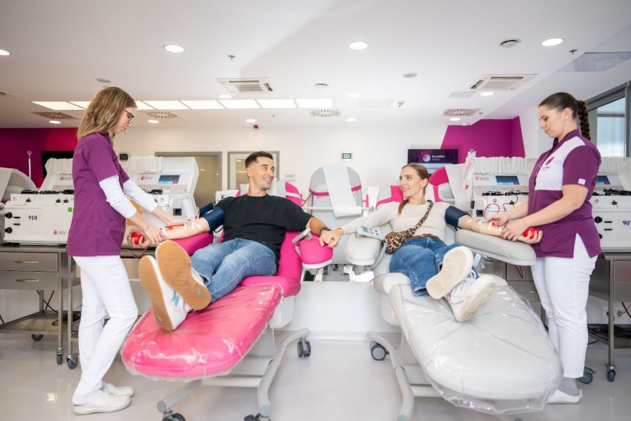 Két fiatal plazmaadó egymás mellett fekszik a plazmaadáson, kezükben piros stresszlabdával, miközben egészségügyi dolgozók segítik a folyamatot.